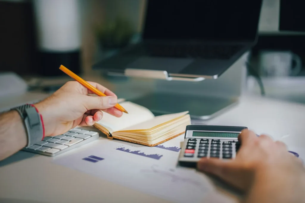 People using a calculator and notebook beside a laptop