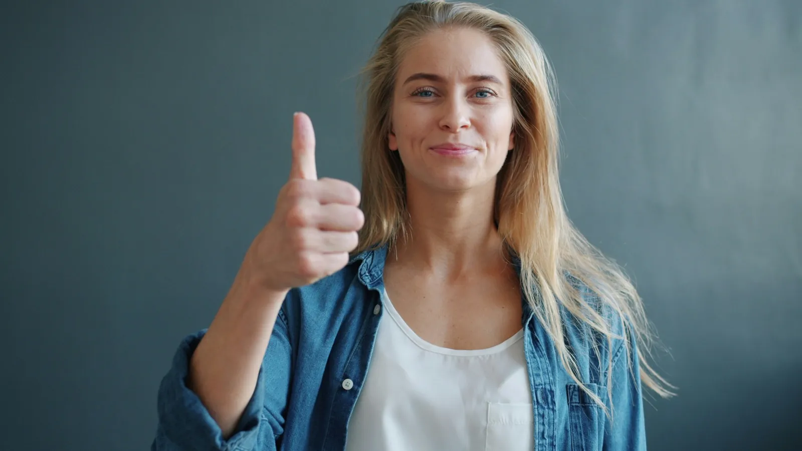 Woman in a denim shirt giving a thumbs-up against a gray background