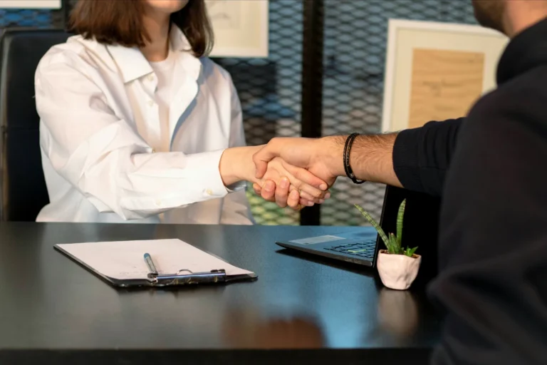 Two people shaking hands across an office desk.